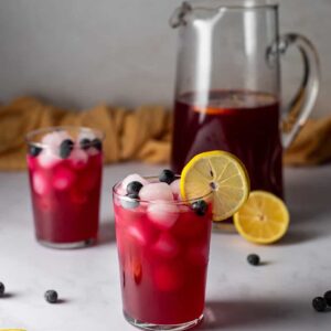 two glasses of blueberry lemonade, a pitcher of blueberry lemonade in the background