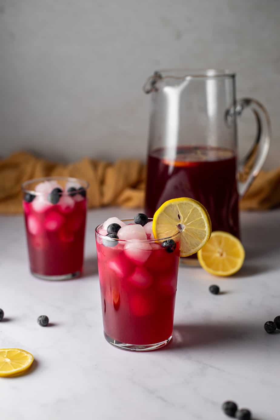 two glasses of blueberry lemonade, a pitcher of blueberry lemonade in the background