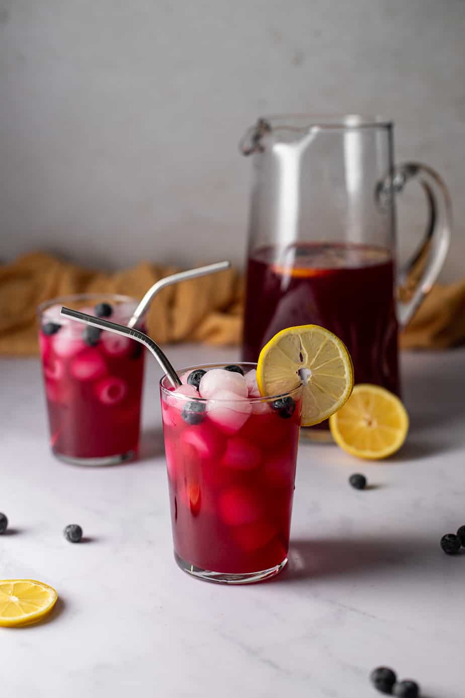 two glasses of blueberry lemonade with metal straws, a pitcher of blueberry lemonade in the background