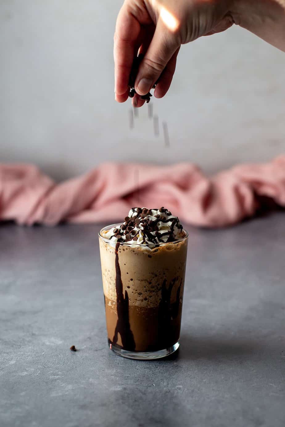 a java chip frappuccino in a glass, a hand is sprinkling mini chocolate chips on top