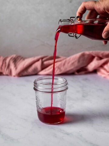 cherry simple syrup being poured from a glass bottle into a glass jar