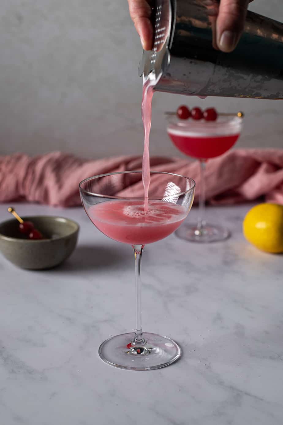 a cocktail shaker pouring a cherry vodka sour into a coupe glass.