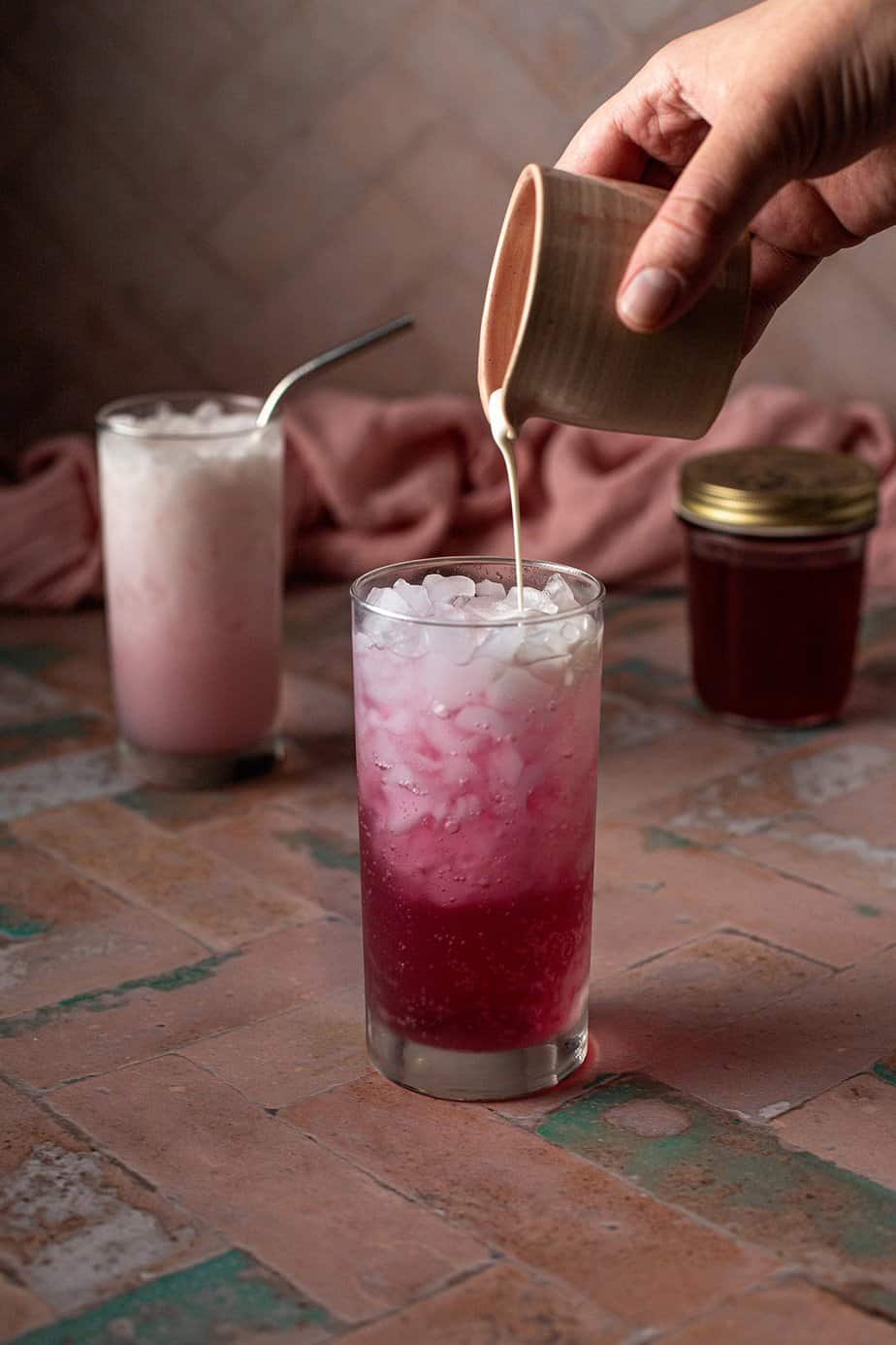 a tall glass with crushed ice, club soda, cherry simple syrup, and cream being poured in