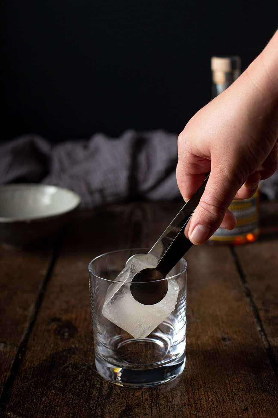 a hand using tongs to set a large ice cube in a rocks glass