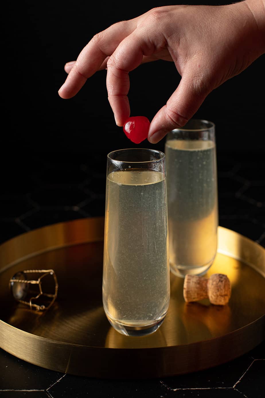 two stemless champagne flutes on a gold serving tray, full of french 76 cocktail, with a hand holding a cherry over one flute