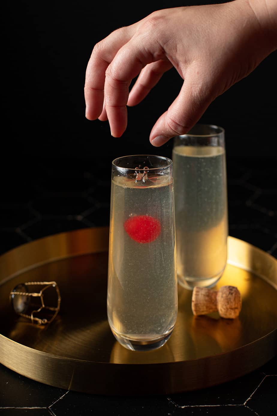 two stemless champagne flutes on a gold serving tray, full of french 76 cocktail, with a hand dropping a cherry into one of the flutes