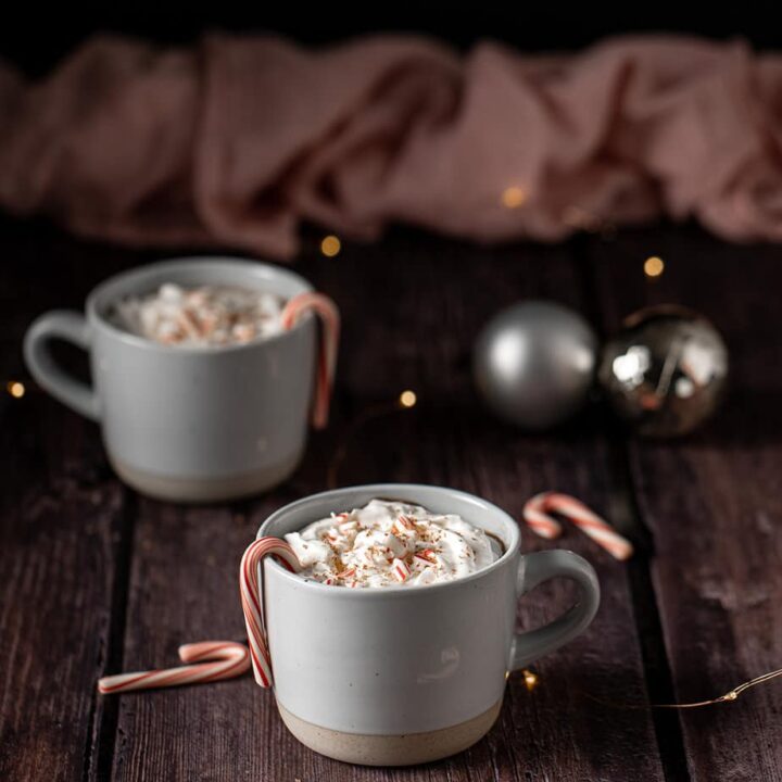 two mugs full of peppermint hot chocolate topped with whipped cream, crushed candy canes, and chocolate shavings