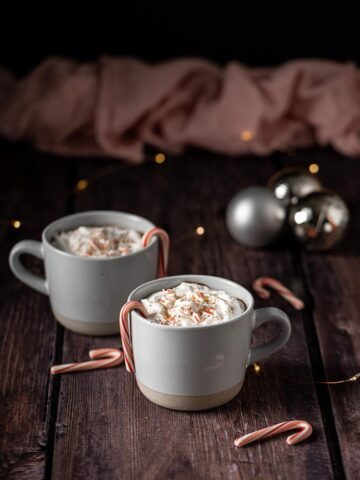 two mugs full of peppermint hot chocolate topped with whipped cream, crushed candy canes, and chocolate shavings