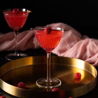 a nick and nora glass with raspberry martini in it on a gold tray