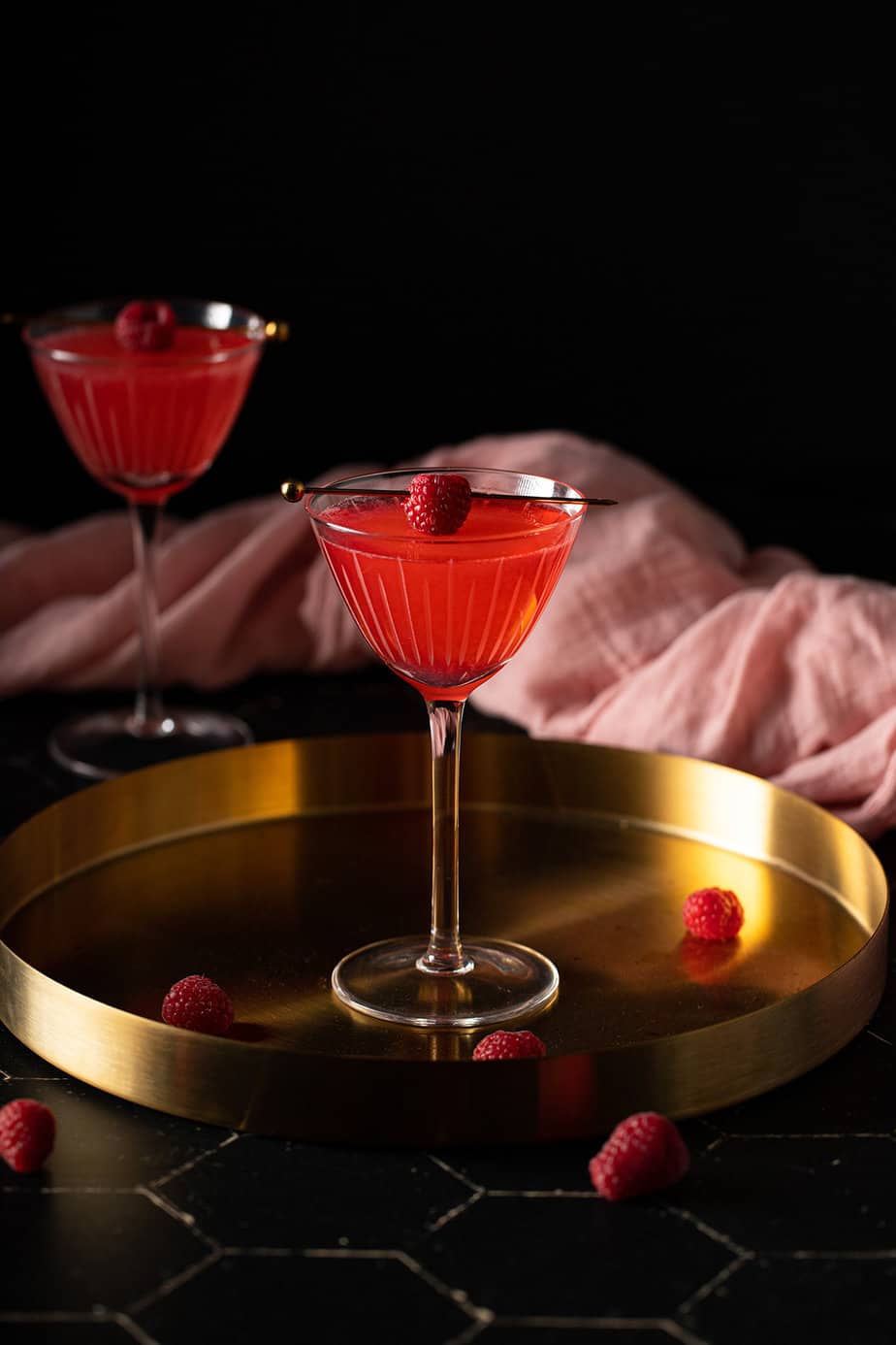 a nick and nora glass with raspberry martini in it on a gold tray