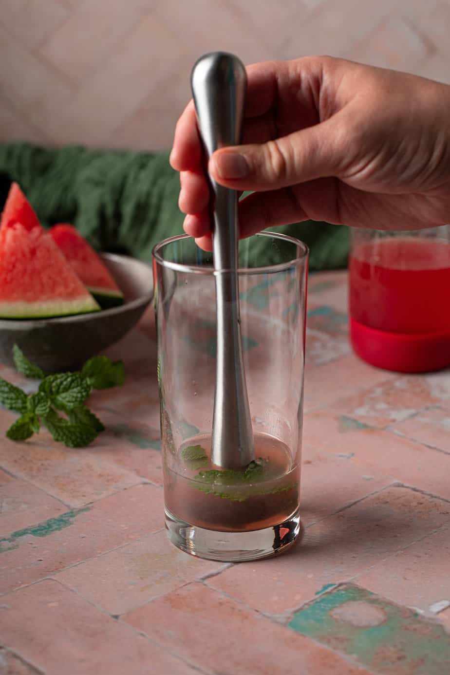 tall glass with mint leaves, watermelon syrup, and lime juice; a cocktail muddler is muddling the ingredients.