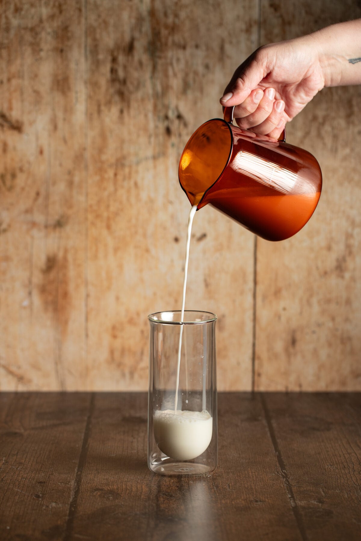 a hand pouring a pitcher of milk into a tall clear glass.