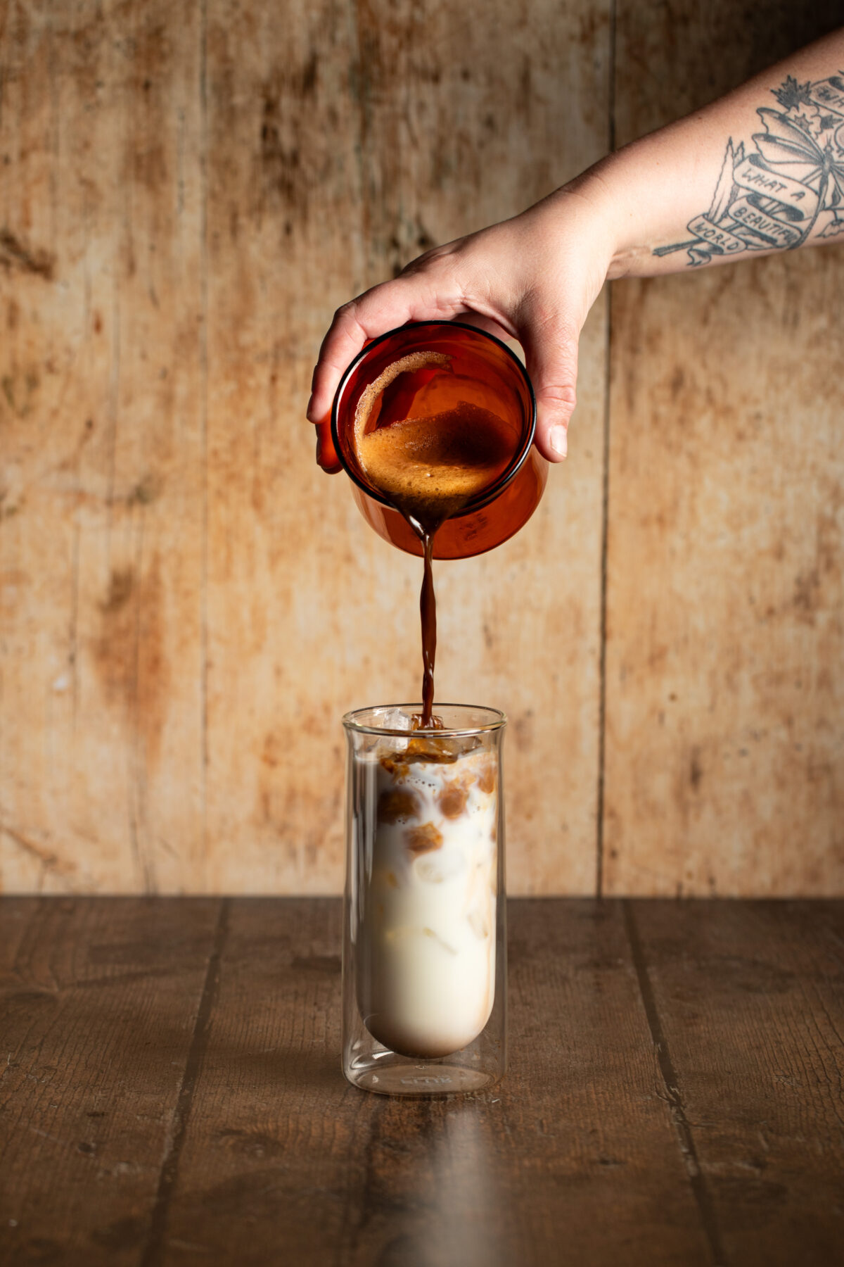 a hand pouring espresso into a tall clear glass full of milk and ice.