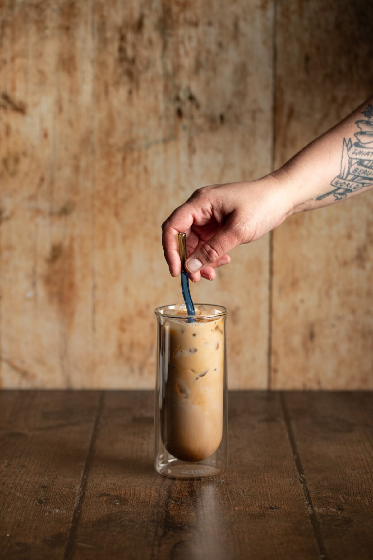 a tall clear glass full of an iced latte, a hand holding a straw is stirring the latte.